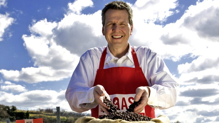 Person in roter Schürze hält Kaffeebohnen über einen offenen Kaffeesack, im Hintergrund rote Verpackungen und eine Plantage.