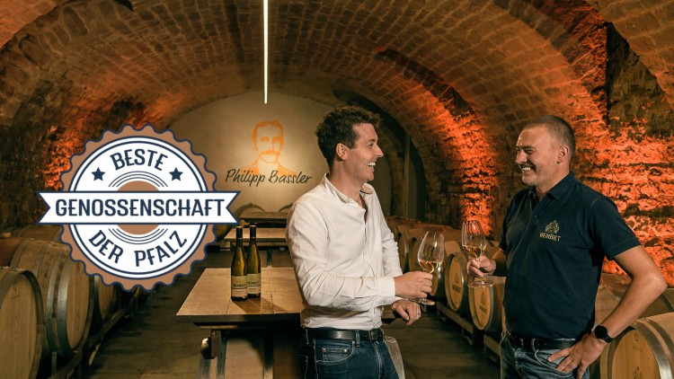 Zwei Personen stehen mit Weingläsern in einem Weinkeller, Holzfässer im Hintergrund, Logo „Beste Genossenschaft der Pfalz“.