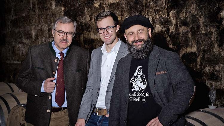 Drei Personen stehen vor einer Wand aus Stein in einem Weinkeller mit mehreren Holzfässern im Hintergrund.