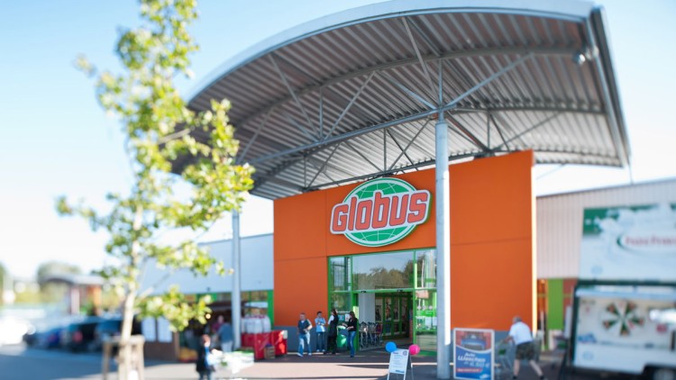 Eingangsbereich eines GLOBUS Marktes mit orangefarbener Fassade, Metallvordach und Kundinnen und Kunden vor dem Eingang.