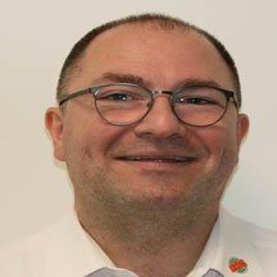 Eine weiße Hemdbluse mit roter und grüner Logo-Dekoration wird vor einem hellen Hintergrund getragen.