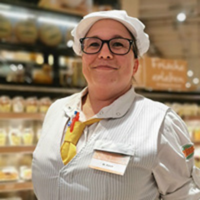 Eine Person in gestreifter Arbeitskleidung mit Namensschild steht vor einem Regal mit verpackten Lebensmitteln.