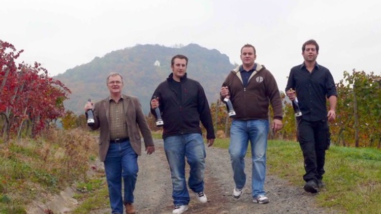 Vier Personen gehen einen Feldweg entlang und tragen Weinflaschen, im Hintergrund sind Hügel und Weinreben zu sehen.
