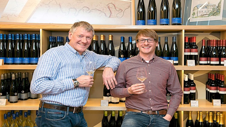Zwei Personen stehen vor Regalen mit Weinflaschen und halten jeweils ein Glas Weißwein in der Hand.