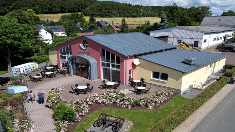 Ein Gebäude mit der Aufschrift „Grüner Hof“, umliegenden Tischen und Stühlen sowie Blumenbeeten, ländliche Umgebung im Hintergrund.