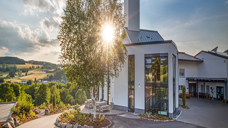 Modernes Gebäude mit weißen Fassaden, großen Fenstern und Terrasse, umgeben von grüner Landschaft, Sonne scheint durch Baum.