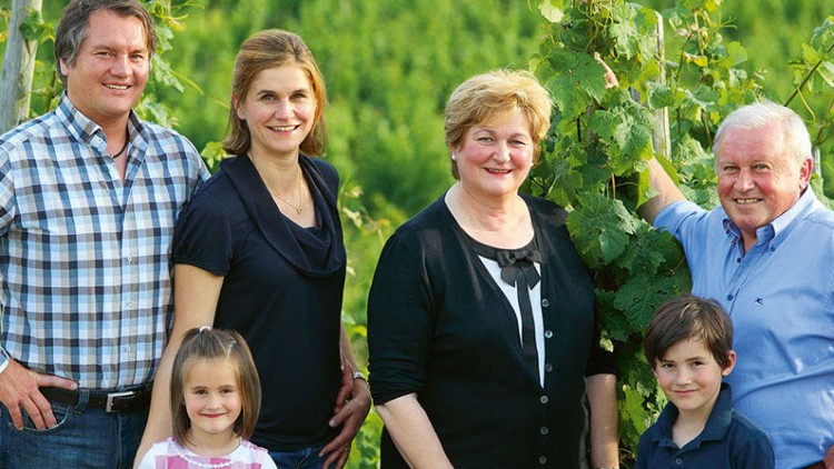 Sechs Personen stehen zusammen vor Weinstöcken in einer grünen Landschaft, umgeben von Pflanzen und Stützpfosten.