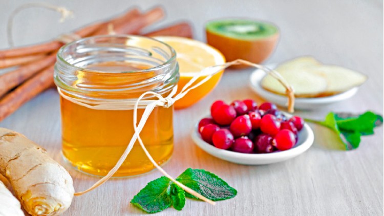 Ein Glas mit Honig, daneben frische Minze, Ingwer, Cranberries, eine halbe Kiwi, Zimtstangen und Orangenhälften.