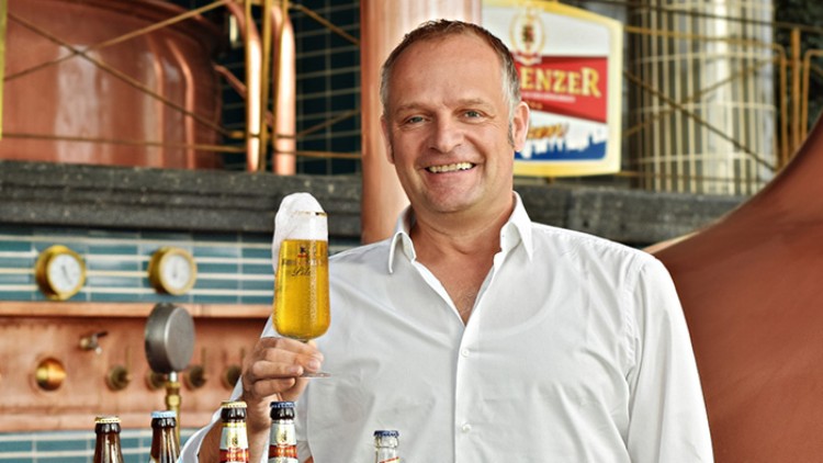 Person in weißem Hemd hält ein Bierglas mit Schaumkrone vor einer Brauereikulisse mit Kupferkesseln und mehreren Bierflaschen.