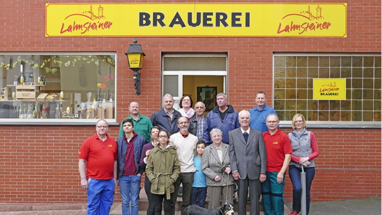 Eine Gruppe von Personen steht vor einem Gebäude mit gelbem Schild und roter Aufschrift „Lahmsteiner BRAUEREI“.