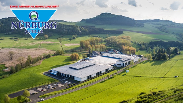 Landschaft mit grünen Feldern und Hügeln, zentral Gebäudeanlage, oben links das Logo „NÜRBURG QUELLE, Das Mineralienwunder“.
