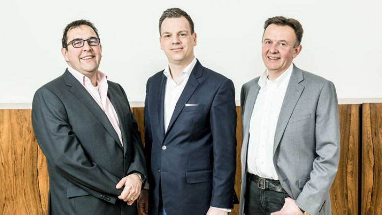 Drei Personen tragen Anzüge und stehen vor einer weißen Wand mit einer Vertäfelung aus Holz.