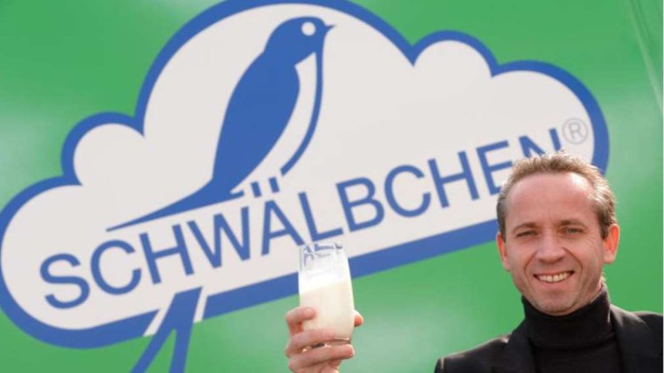 Ein Glas Milch wird vor einem grünen Hintergrund mit dem blauen Logo und Schriftzug „SCHWÄLBCHEN“ hochgehalten.