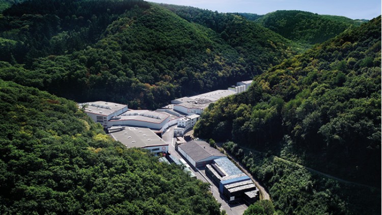 Industrieanlage mit mehreren großen Gebäuden, umgeben von dicht bewaldeten Hügeln und grüner Landschaft.