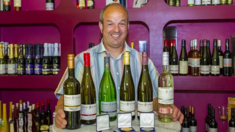 Sechs Weinflaschen mit verschiedenen Farben und Etiketten sowie drei Medaillen stehen auf einem runden Tisch vor einem Weinregal.