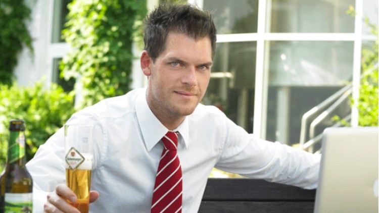 Person in weißem Hemd und roter Krawatte sitzt draußen, hält ein Bierglas, im Hintergrund Grünpflanzen und Fenster.