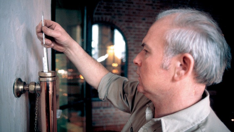 Person hält ein Thermometer in einem kupferfarbenen Rohr vor einer Wand, Hintergrund mit Ziegelmuster und Fenstern.
