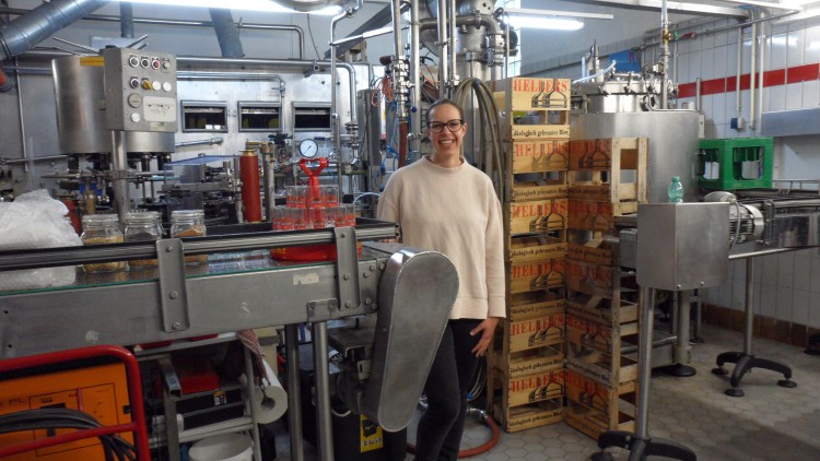 Produktionshalle mit Förderbändern, Abfüllanlage, Metallrohren und gestapelten Bierkisten der Sorte Hellas im Hintergrund.