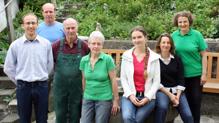Sieben Personen stehen oder sitzen vor einer grünen Gartenkulisse mit Bank, Treppe und Zierfigur.