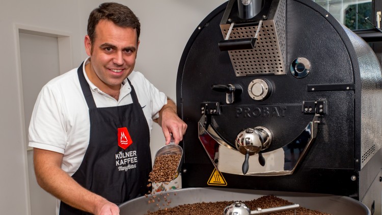 Person in dunkler Schürze mit Aufschrift „Kölner Kaffee Manufaktur“ schüttet Kaffeebohnen in eine silberne Röstanlage.