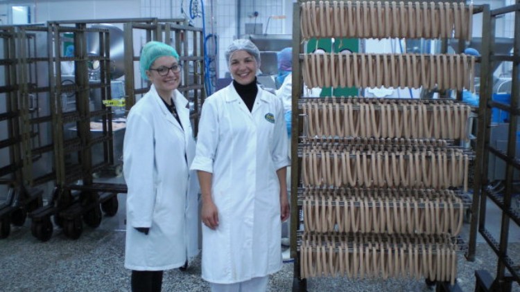 Zwei Personen in weißer Arbeitskleidung stehen in einer Produktionshalle neben einem Gestell mit hängenden Würsten.