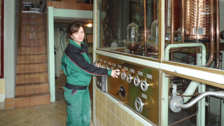 Person in grüner Arbeitskleidung bedient eine Schalttafel vor kupfernen Destillationsapparaten in einem technischen Raum.