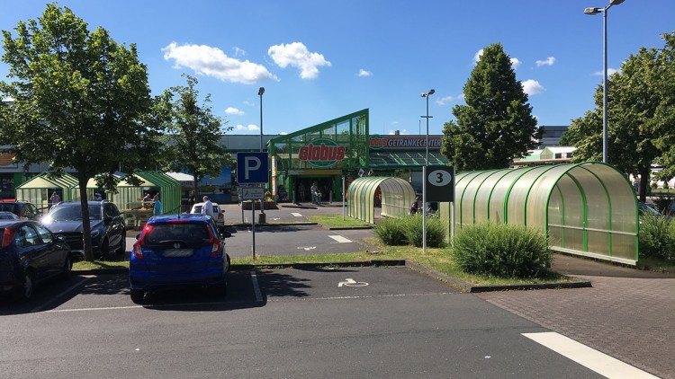 Parkplatz vor einem GLOBUS-Markt mit grünen Einkaufswagen-Unterständen, Autos, Bäumen und blauem Himmel.