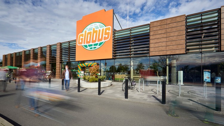 Eingang eines GLOBUS Marktes mit orangenem Logo, Glasfassade, Fahrradständern und Personen auf dem Vorplatz.