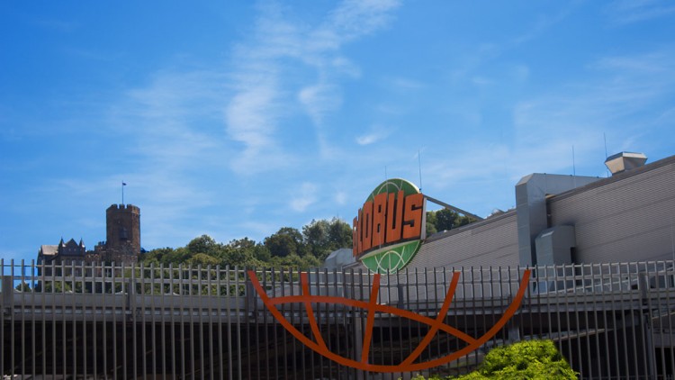 Ein GLOBUS-Schild mit orange-grünem Logo vor einem Gebäude, im Hintergrund eine Burg und grüne Baumkronen unter blauem Himmel.