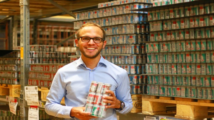 Mann in hellblauem Hemd hält ein Sixpack Dosen vor einem Regal mit gestapelten Dosenpaletten in einem Lager.