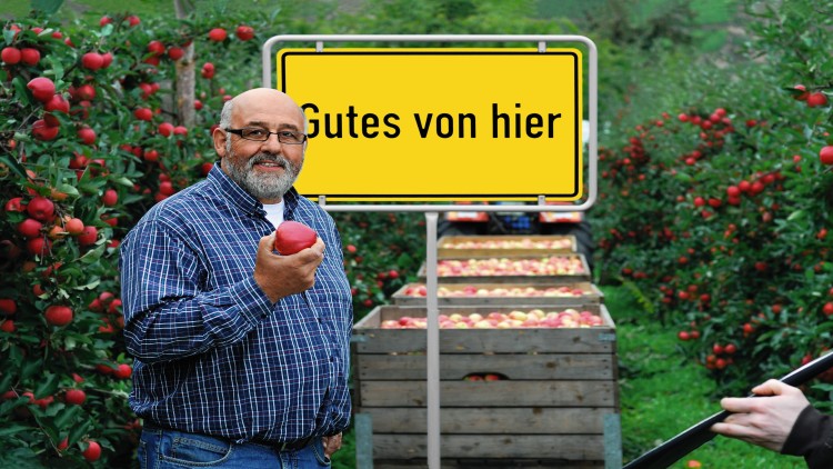 Ein Apfelbauer hält einen roten Apfel, im Hintergrund Kisten mit Äpfeln, Apfelbäume und ein gelbes Schild mit „Gutes von hier“.