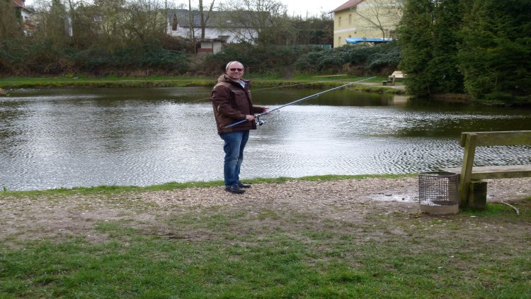 Person angelt mit einer blauen Angelrute an einem Teich, im Hintergrund Bäume und Gebäude, daneben grüne Wiese und Mülleimer.
