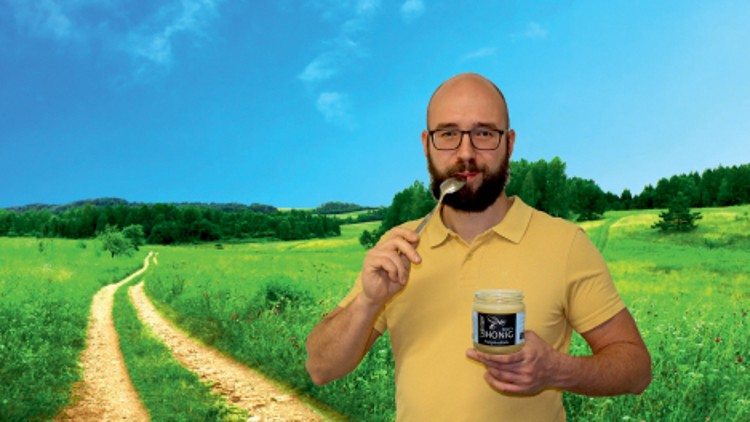 Person in gelbem Polohemd hält ein Glas mit der Aufschrift „Honig“ in der Hand, im Hintergrund eine grüne Wiesenlandschaft.