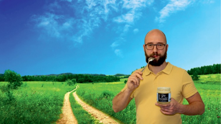 Person in gelbem Poloshirt hält ein Honigglas in Landschaft mit grünem Feld, Weg und wolkenlosem Himmel.