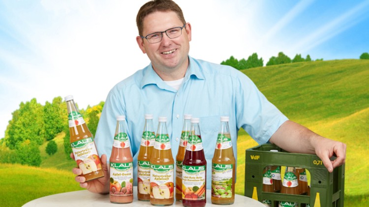 Sechs Flaschen KLASS Apfelsaftgetränke stehen auf einem Tisch, daneben eine grüne Getränkekiste vor Landschaft.