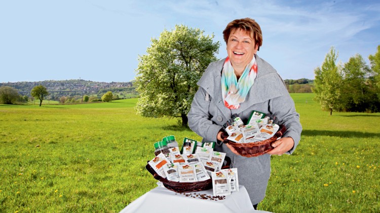 Person hält zwei Körbe mit verpackten Lebensmitteln vor einer Wiese mit Bäumen und blauem Himmel.