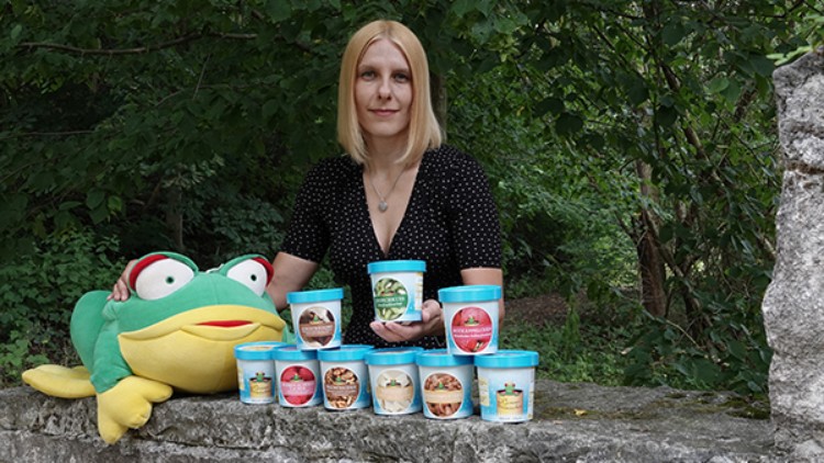 Eine Person hält Eisbecher, weitere Becher stehen auf einer Steinmauer neben einem großen Stofffrosch, Hintergrund mit Bäumen.