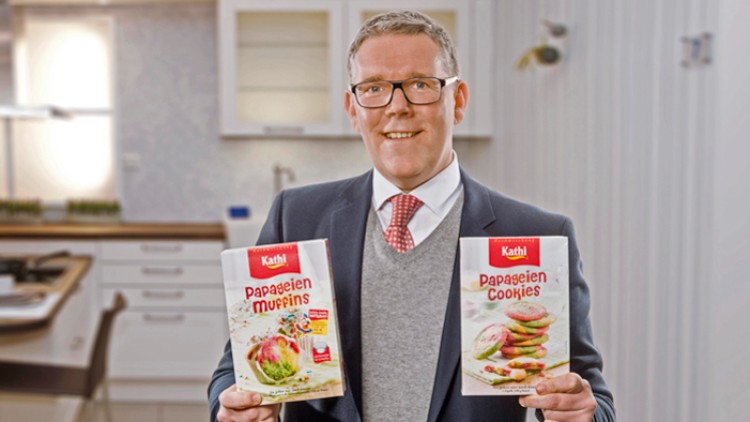 Person hält in einer Küche zwei Packungen hoch, eine mit der Aufschrift „Papageien Muffins“ und die andere „Papageien Cookies“.