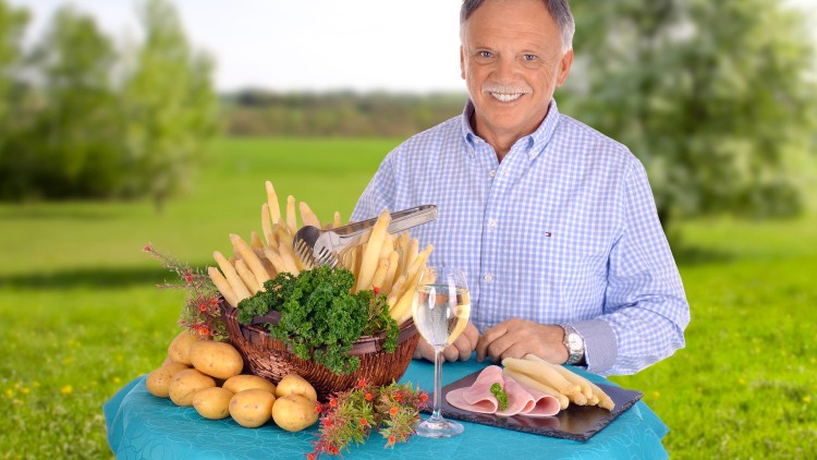 Ein Korb mit weißem Spargel, Petersilie und Kartoffeln, daneben ein Teller mit Spargel und Schinken sowie ein Glas Weißwein.