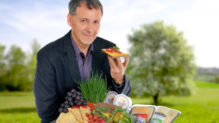 Ein rechteckiger Tisch mit Käseplatte, Gemüse, Brot, Trauben, Frischkäse-Produkten und einer Wiese mit Bäumen im Hintergrund.