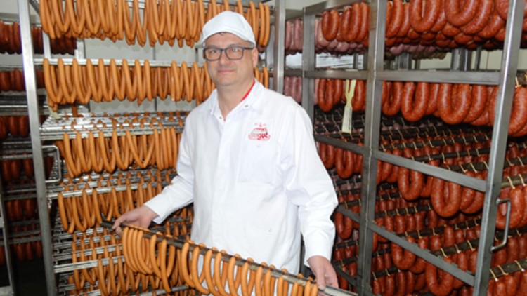 Person in weißer Arbeitskleidung hält Wurststange vor Regalen mit hängenden Würsten in einer Fleischverarbeitung.