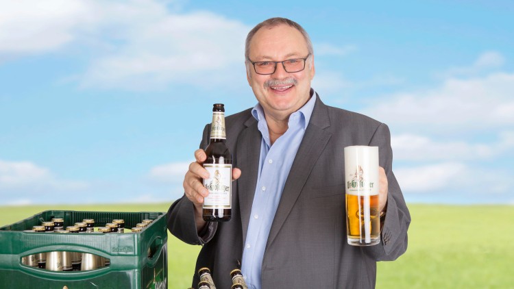 Person in Anzug hält eine Flasche Ur-Krostitzer Bier und ein gefülltes Bierglas, daneben grüne Bierkästen vor Wiese.