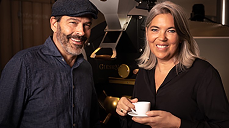 Eine Person hält eine weiße Tasse, im Hintergrund ist eine große schwarze Kaffeemaschine mit goldenen Akzenten zu sehen.