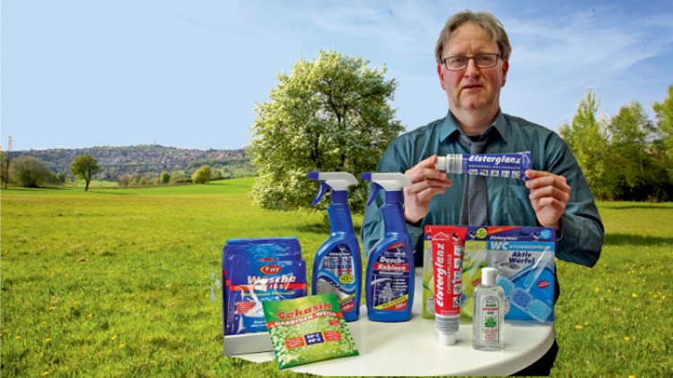 Mehrere Putz- und Reinigungsprodukte wie Sprays, Pulver und Gel stehen auf einem weißen Tisch vor einer grünen Landschaft.