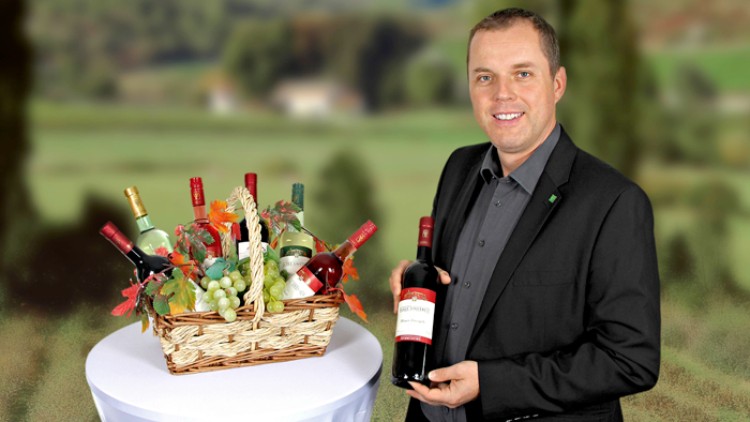 Ein geflochtener Korb mit Weinflaschen, Trauben und Dekoration steht auf einem weißen Tisch, daneben eine Person mit Weinflasche.