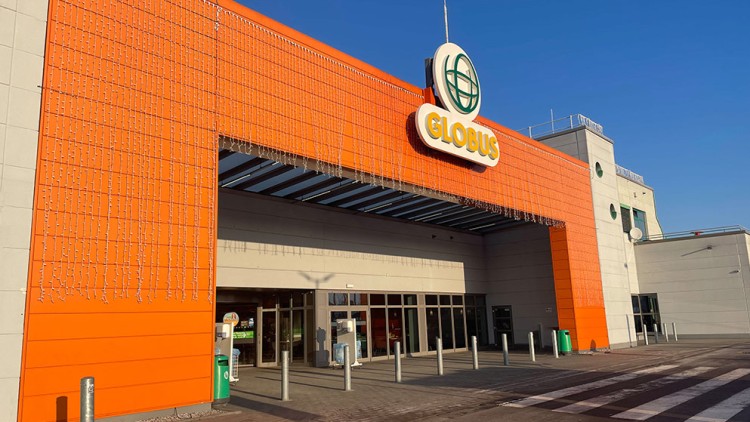 Eingangsbereich eines GLOBUS Gebäudes mit oranger Fassade, beleuchtetem Logo, Glasdach und Eiszapfen-Lichterketten.