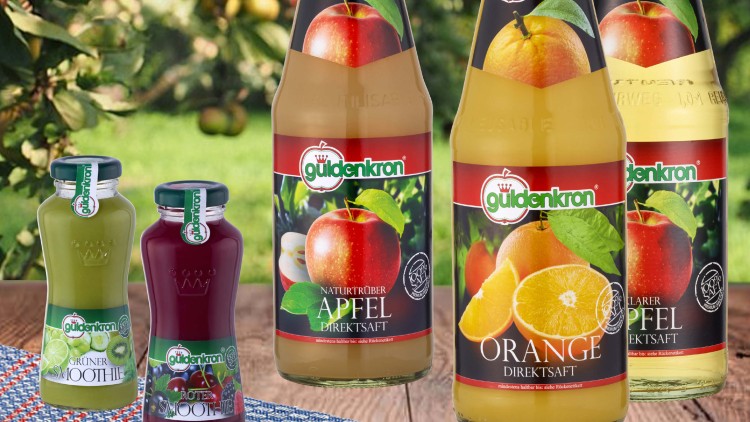 Verschiedene Flaschen von Güldenkron-Säften und Smoothies stehen vor einer Obstbaumwiese, Etiketten zeigen Sorten wie Apfel und Orange.