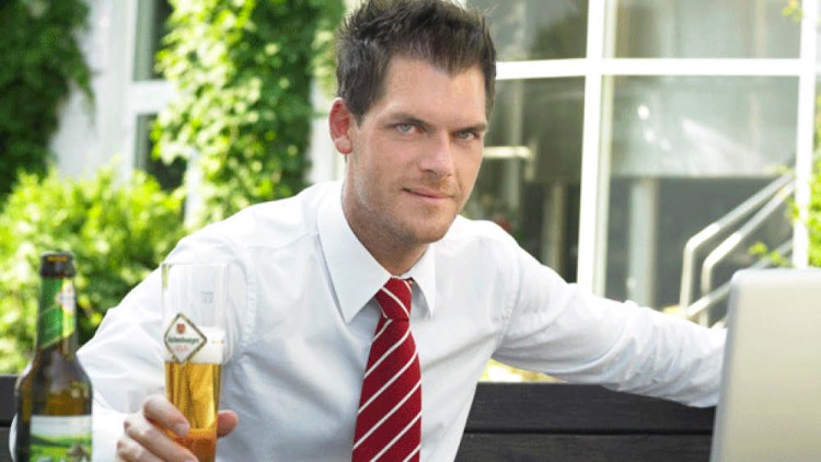Person in weißem Hemd und roter Krawatte hält ein Glas Bier, daneben steht eine Bierflasche auf einem Tisch im Freien.