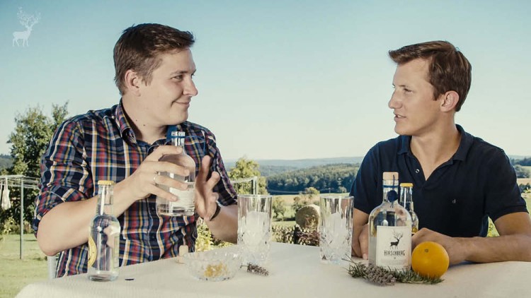 Zwei Personen sitzen an einem Tisch mit Gläsern, einer Flasche Hirschberg-Getränk, einem Orangenstück und Zweigen, im Freien.