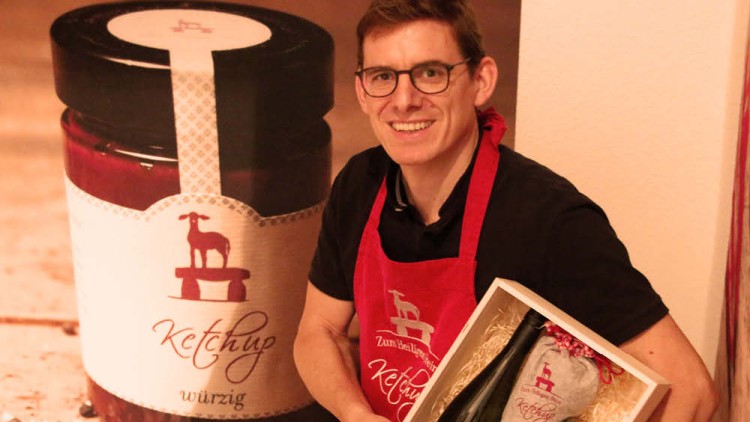 Ein großes Ketchupglas mit rotem Inhalt und Ziegenlogo, daneben eine Person mit roter Schürze und Geschenkbox mit Flasche.